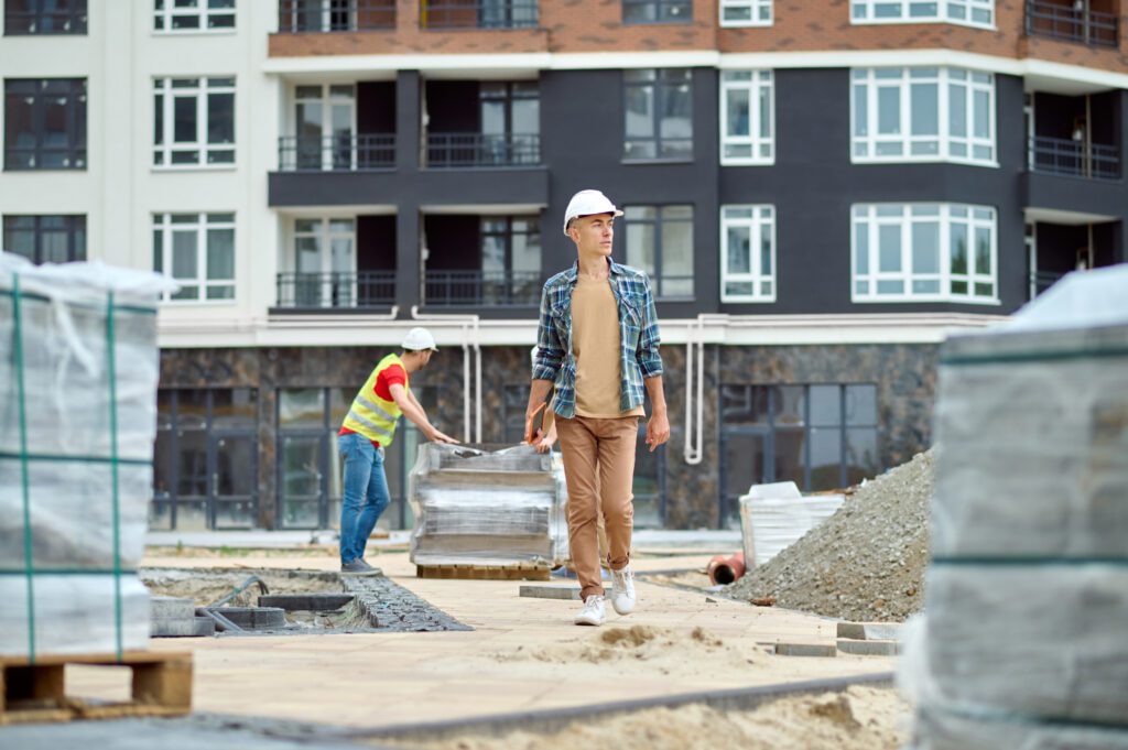Control. Man in protective helmet with tablet walking on construction site looking to side and worker standing behind near building materials in afternoon