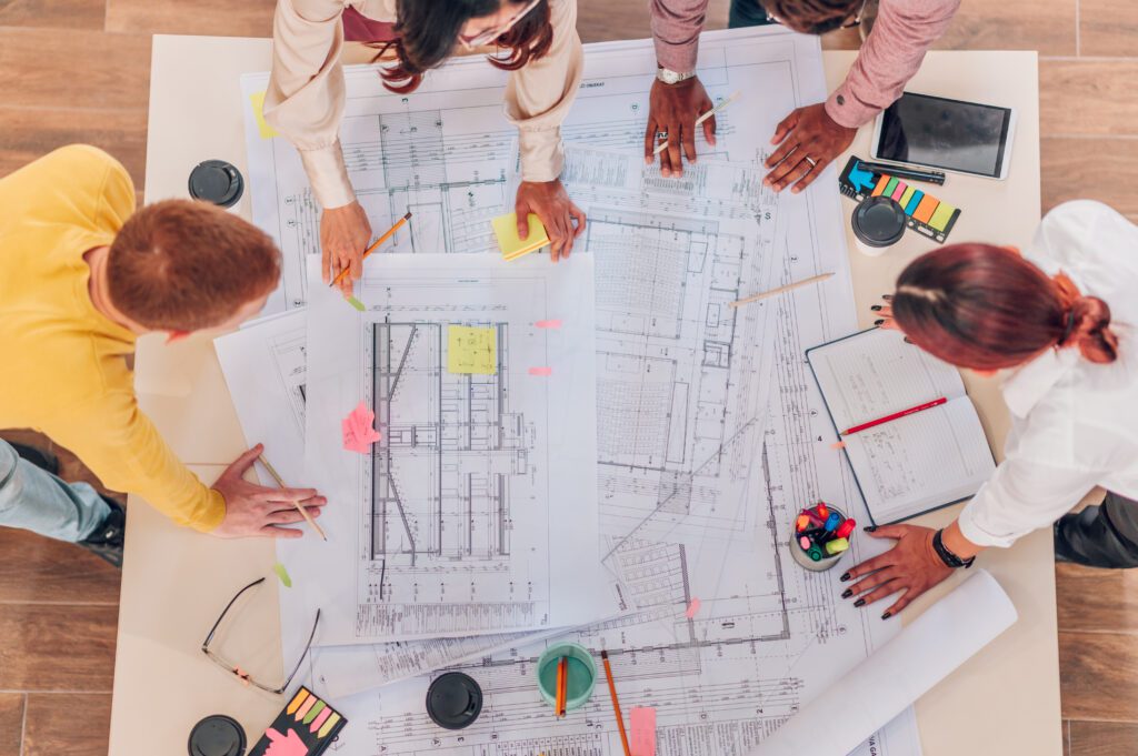 Cropped shot of a multiracial group of architects gathering at table with blueprints and gadgets while taking notes about new project they are working on. Focus on a construction plans.