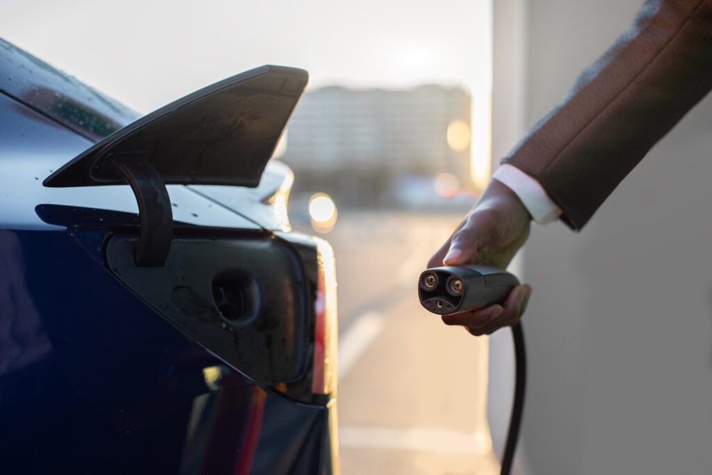 Close up cropped image of unrecognizable afro american man in business wear, holding electricity cable for electric vehicle charging man's hand preparing to charge an electric car at outdoor charging station in the city